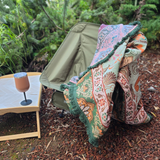 Vanaway Willow woven travel throw rug featuring soft blue, sage green, cream and terracotta botanical patterns, draped over a camping chair with decorative green fringe in a bush camping setting.